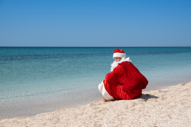 Papai Noel com um saco de presentes sentado na praia. Céu azul. Água turquesa. Copie o espaço para o seu texto - Imagem