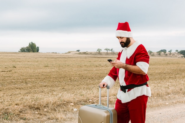 Papai Noel com uma mala está verificando um telefone celular. Conceito de férias de natal