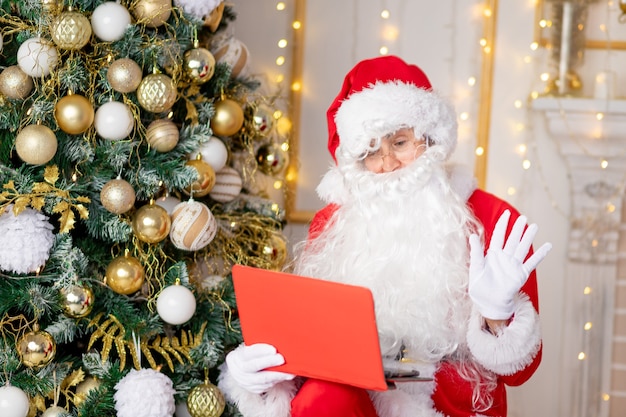 Papai Noel em casa perto da árvore de natal com um tablet parabeniza por vídeo link, o conceito de ano novo e natal