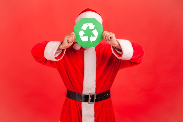 Papai Noel em pé cobrindo o rosto com a placa verde de reciclagem, meio ambiente, ecologia.