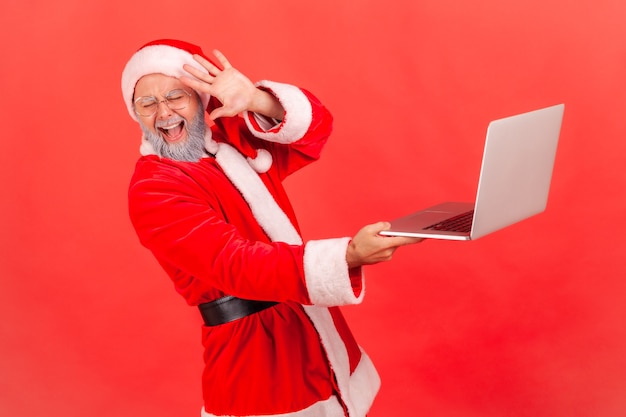 Papai Noel em pé com o computador nas mãos, vê um conteúdo terrível na tela, cobrindo o rosto.