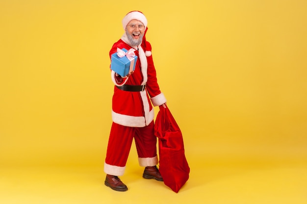 Foto papai noel em pé com um saco vermelho com presentes e segurando uma caixa de presente de natal.