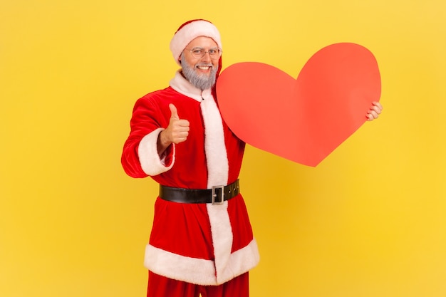 Papai noel em pé segurando um grande coração vermelho na mão, mostrando o polegar para a câmera com um sorriso.