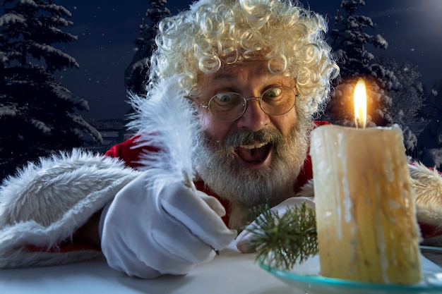 Papai Noel emocional parabenizando com o ano novo de 2021 e o Natal. Homem em traje tradicional, escrevendo uma carta, lista de desejos iluminada com velas e floresta à noite no fundo. Férias de inverno.