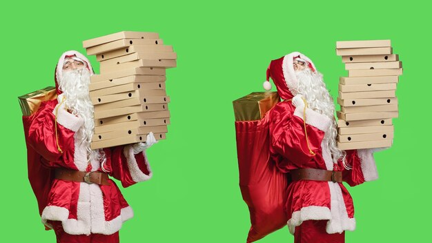 Foto papai noel entregando caixas de pizza em tela verde, carregando uma grande pilha de fast food e saco com brinquedos para crianças. são nick em traje vermelho festivo agindo como um entregador em estúdio.