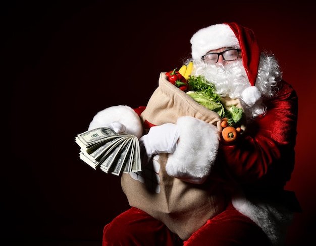 Papai Noel está segurando uma grande sacola cheia de frutas e vegetais em um pacote de 100 dólares de