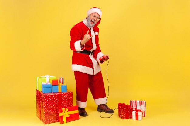 Papai noel jogando videogame, em pé com um joystick, mostrando o sinal de pedra para a câmera.