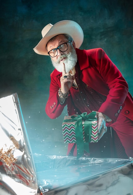 Papai Noel moderno e elegante em um terno vermelho da moda e chapéu de cowboy na parede escura