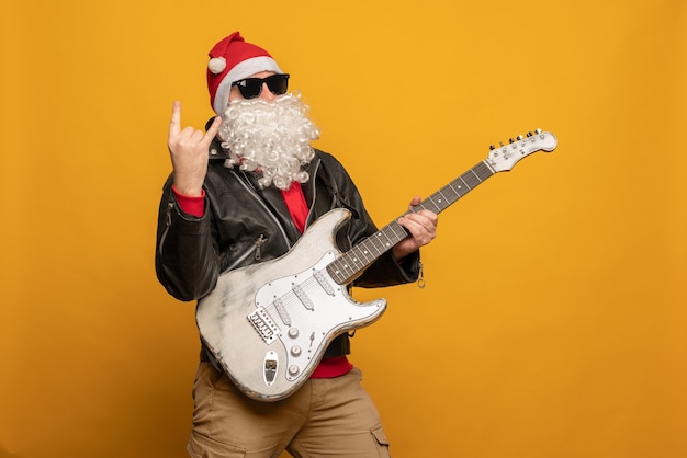 Papai Noel moderno em jaqueta de couro, rebelde rock n roller toca guitarra emocionalmente isolado em fundo amarelo