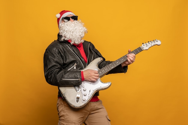 Papai Noel moderno em jaqueta de couro, rebelde rock n roller toca guitarra emocionalmente isolado em fundo amarelo