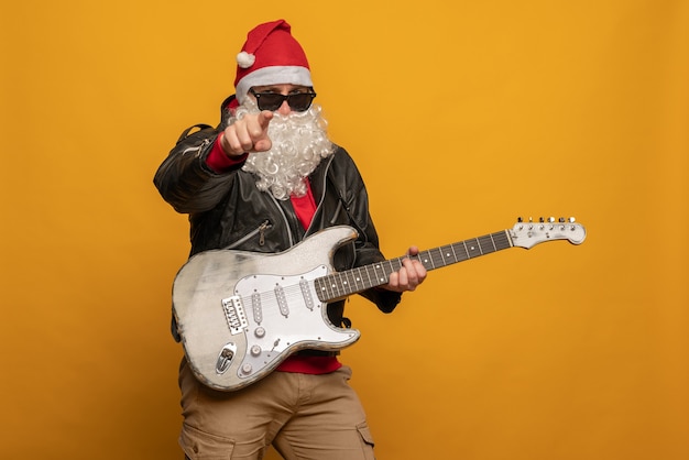 Papai Noel moderno em jaqueta de couro, rebelde rock n roller toca guitarra emocionalmente isolado em fundo amarelo