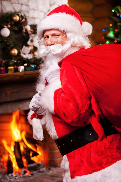 Papai noel. papai noel tradicional carregando um saco com presentes e sorrindo com uma árvore de natal e uma lareira ao fundo