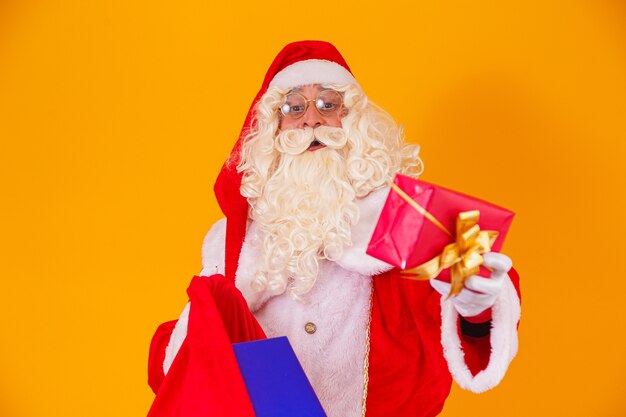 Papai Noel real em fundo amarelo segurando o saco com vários presentes de Natal.