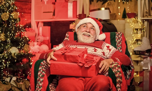 Papai Noel relaxando na poltrona Homem sênior barbudo Papai Noel Lenda sobre Papai Noel Feliz natal Entregando presentes Presentes para a família Conceito de tradições Vovô idoso em casa
