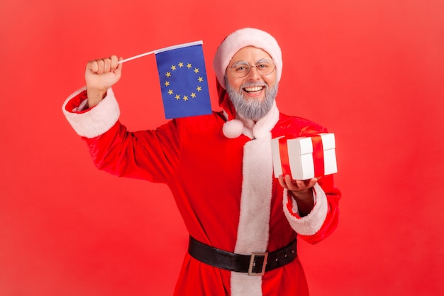 Papai Noel segurando a caixa de presente embrulhada e acenando a bandeira da União Europeia, olha para a câmera.