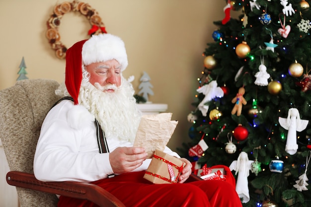 Papai noel sentado com cartas de crianças em uma cadeira confortável perto da lareira em casa