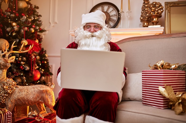 Papai noel sentado em sua casa lendo e-mail no laptop com ñ pedidos de natal ou lista de desejos perto da lareira e árvore com presentes. ano novo e feliz natal, conceito de boas festas