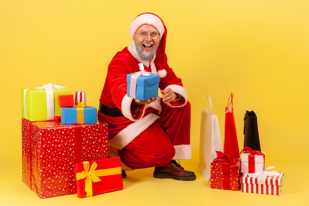 Foto papai noel sentado perto de caixas de presentes embrulhadas e dando um presente, parabenizando com o ano novo.