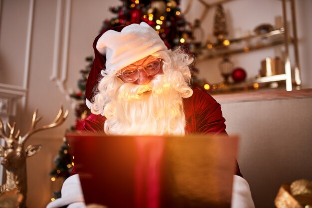 Papai Noel surpreso com um presente mágico e brilhante perto da bela árvore de Natal. Feliz Natal, conceito de boas festas