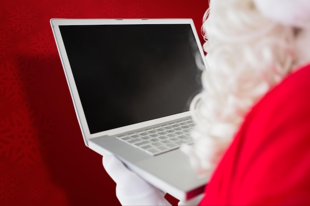 Foto papai noel usando seu laptop contra o fundo vermelho do floco de neve