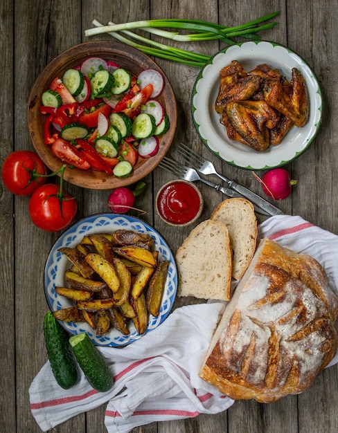 Papas fritas, alitas, ensalada, verduras, pan sobre un fondo de madera vieja. Cena rural, picnic de verano. Vista superior. Endecha plana.