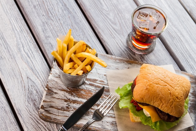 Papas fritas y hamburguesa. Cubiertos y vaso con cola. Comida apetitosa en la cafetería. Es hora de cenar.