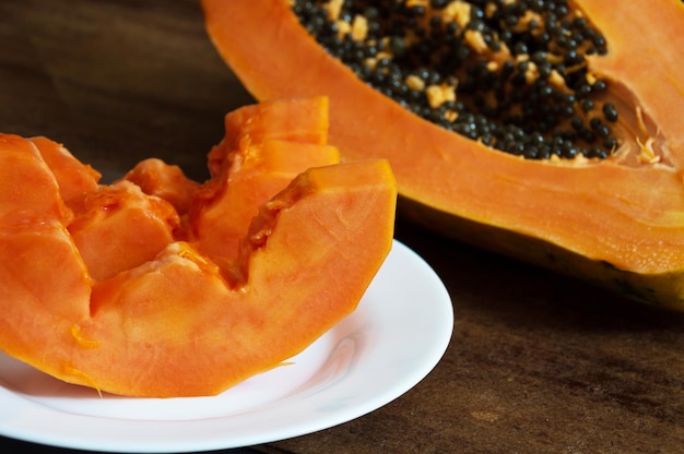 Foto papaya en rodajas en un plato blanco y media papaya con semillas ligeramente fuera de foco en la esquina superior derecha