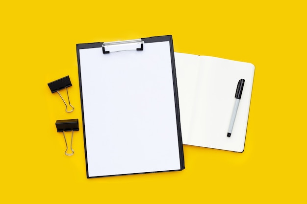 Papel a4 en blanco en el portapapeles negro, clips de oficina negros con cuaderno y bolígrafo sobre fondo amarillo.