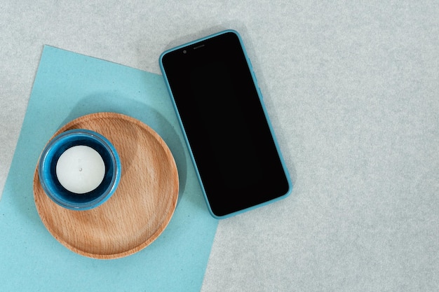 Papel azul de teléfono inteligente en blanco con vela y plato de madera sobre fondo gris