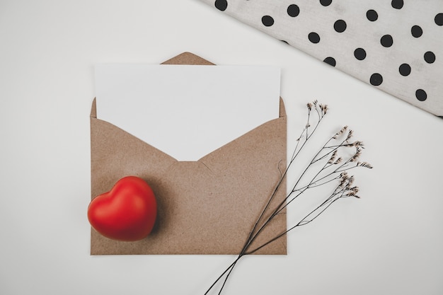 Papel branco em branco é colocado em envelope de papel marrom aberto com coração vermelho e flor seca de Limônio