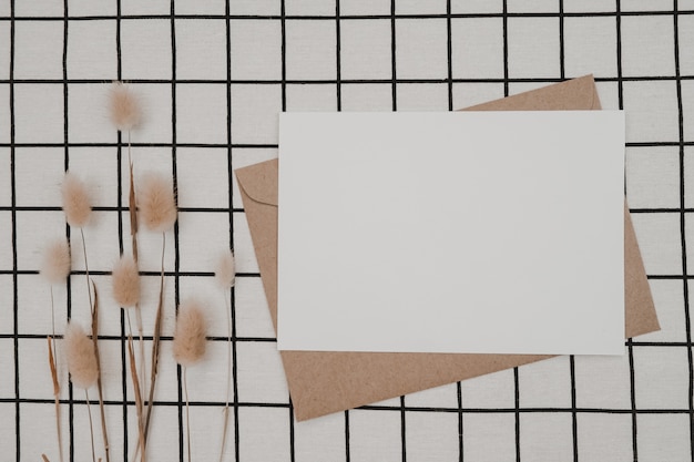 Papel branco em branco em envelope de papel pardo com flor seca de cauda de coelho e caixa de papelão em pano preto com padrão de grade preto branco. mock-up de cartão horizontal em branco.