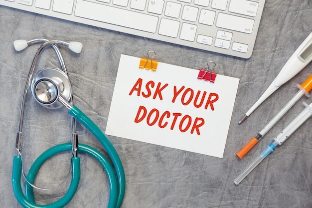 Papel com PERGUNTE A SEU MÉDICO no estetoscópio de mesa de escritório e vista superior do teclado