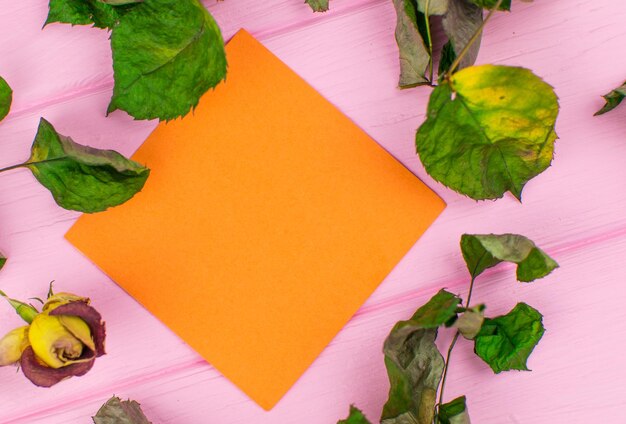 Papel para notas sobre um fundo rosa de madeira com uma flor seca e folhas de rosa mockup