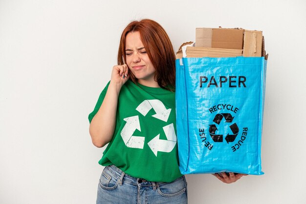 Papel reciclado de la mujer caucásica joven aislado en el fondo blanco que cubre las orejas con las manos.