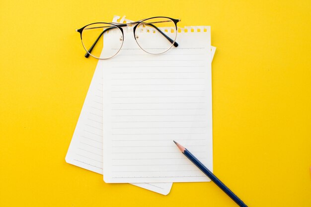 Papel vazio de óculos e lápis do caderno no fundo amarelo com conceito da escola.