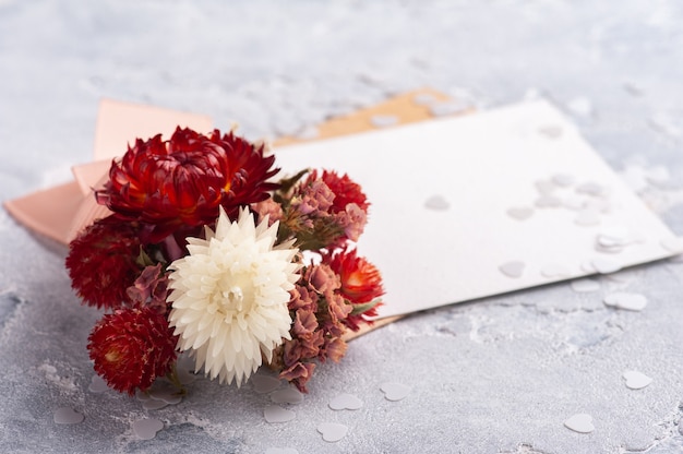 Papel vazio e envelope kraft com buquê vermelho de flores secas. Simulação de casamento na mesa cinza