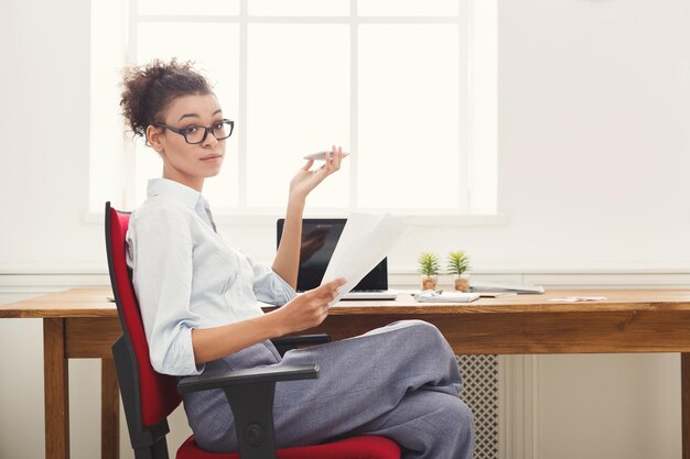 Papelada. Mulher de negócios sérios com roupa formal, sentada em uma mesa de madeira em um escritório moderno, lendo o documento do relatório, vista lateral, copie o espaço