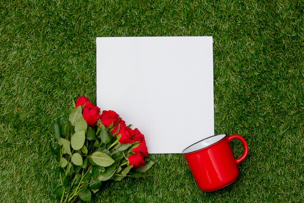 Papelão quadrado branco e buquê de rosas com uma xícara na grama verde.