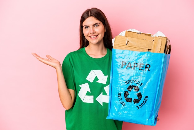 Papelão reciclado jovem mulher caucasiana, isolado no fundo rosa, mostrando um espaço de cópia na palma da mão e segurando a outra mão na cintura.