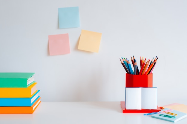 Papelería de colores, lápices de colores y libros brillantes sobre una pared blanca.