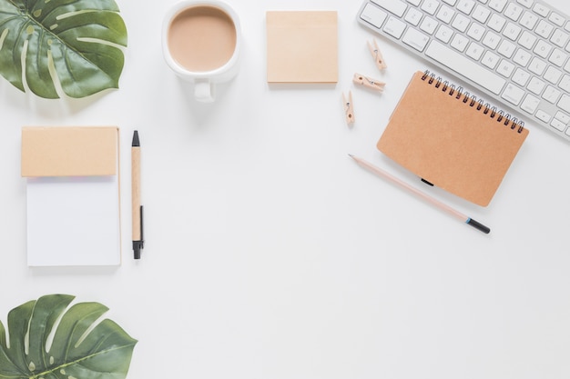 Papelería y teclado en mesa blanca con hojas verdes y taza de café