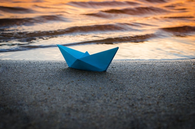 Papierblaues Schiff wird bei Sonnenuntergang auf das sandige Ufer des Sees geworfen.