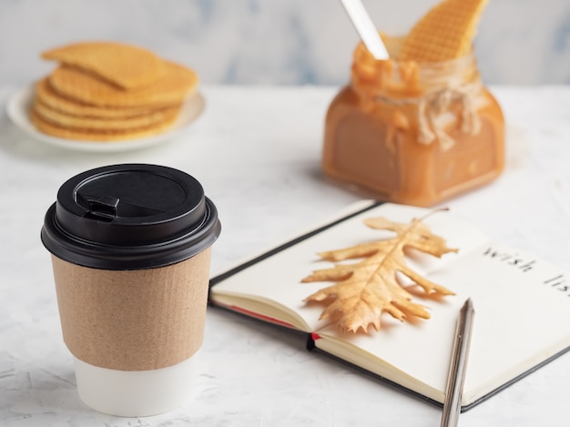 Pappbecher mit Kaffee oder heißem Getränk auf dem Tisch. Notizblock mit einem Wunschzettel. Nahansicht