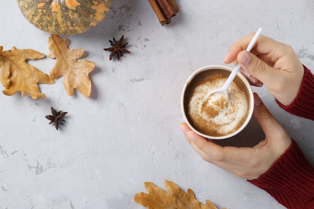 Pappbecher mit köstlichem Kürbislatte mit Gewürzen in den weiblichen Händen auf einem grauen Tisch