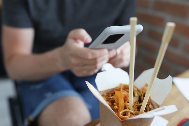 Pappschachtel mit chinesischen Nudeln, die vor dem Hintergrund des Mannes mit dem Handy in der Handnahaufnahme stehen