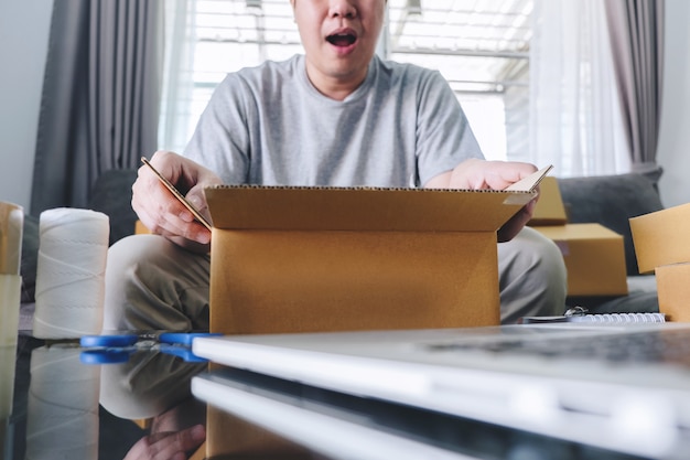 Paquete de la pequeña empresa para el envío, hombre feliz que abre la caja del paquete de las compras en línea con el paquete mientras está sentado en el sofá en casa