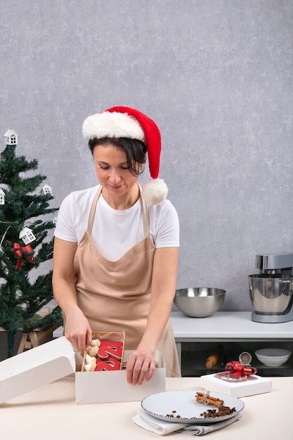 Paquetes de repostería Caja de regalo con sabrosos dulces navideños. Retrato de mujer con sombrero de año nuevo en la cocina. Marco vertical.