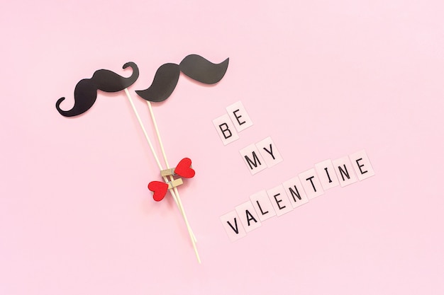 Un par de accesorios de bigote de papel en un palo, una pinza, un corazón y un texto, Be my Valentine