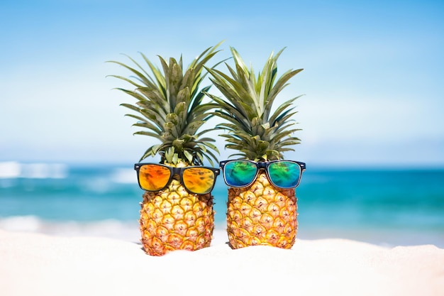 Un par de atractivas piñas con elegantes gafas de sol espejadas en la arena contra el mar turquesa