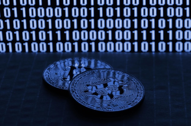Un par de bitcoins se encuentran sobre una superficie de cartón en el fondo de un monitor que representa un código binario de ceros brillantes y una unidad sobre un fondo negro fantasma de color azul clásico